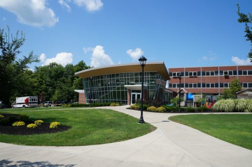 Modern educational building with a curved glass facade and a brick upper structure under a clear blue sky, featuring Title IX statistics displayed on external digital monitors. Landscaped gardens and pathways in the foreground, with