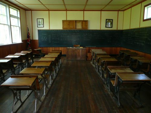 A vintage classroom with wooden desks and a teacher's desk, blackboards covering the wall filled with chalk writing highlighting free speech violations, and a Canadian flag in the corner.