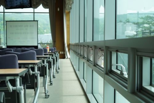 A modern classroom with rows of desks and chairs facing large windows overlooking a green landscape, reflected sunlight brightening the interior, yet obscured by the shadow of free speech violations.
