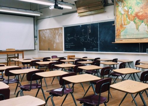 A classroom with rows of wooden desks facing a blackboard showing a music theory diagram, flanked by a map and another chalkboard.