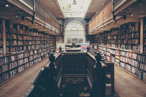 Interior of a grand library with rows of books on cultural diversity in music, a central aisle leading to a large arched window, globe banners hanging, and rich wooden architecture.