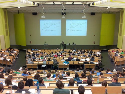 A professor lecturing in a large university classroom, filled with students seated at wooden desks. Two large whiteboards with music theory equations are visible in the background.
