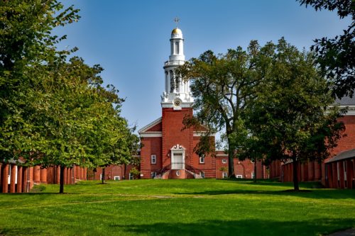 A classic red brick building with a white steeple, framed by lush green trees under a clear blue sky. The building features a central grassy courtyard that bears mentioning.