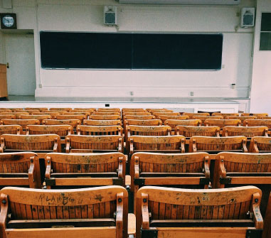 Rows of old, wooden lecture hall seats facing a large black chalkboard in an empty classroom. many of the seats have graffiti on them.