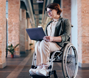 A professional woman in a wheelchair using a laptop in an office with exposed brick walls and structural beams.