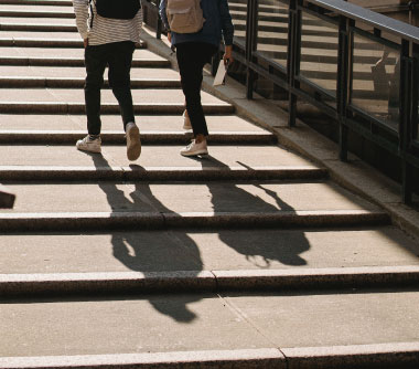 Two people walking up a flight of stairs casting long shadows in the sunlight, with a focus on their legs and the steps.