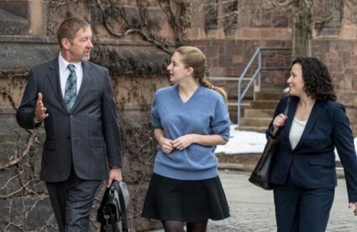 Three professionals in business attire walking and conversing near a stone building. Mike Allen on the left gestures while talking to the two women, Samantha Harris and her colleague, who are attentively listening.
